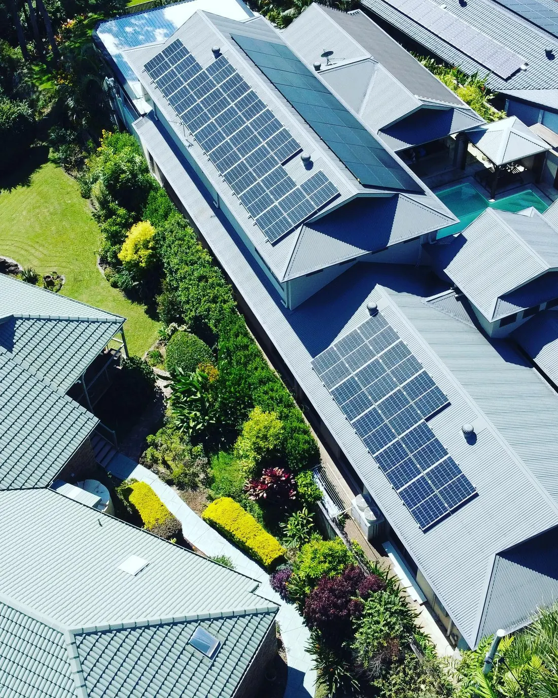 Aerial view of solar panels across several roofs of a coastal home surrounded by gardens