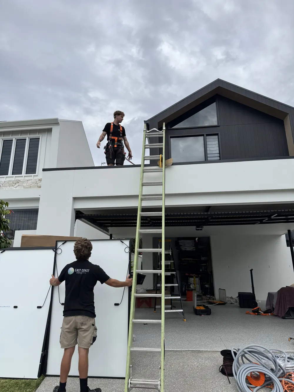 Two East Coast Energy installers moving solar panels up a ladder to a modern home